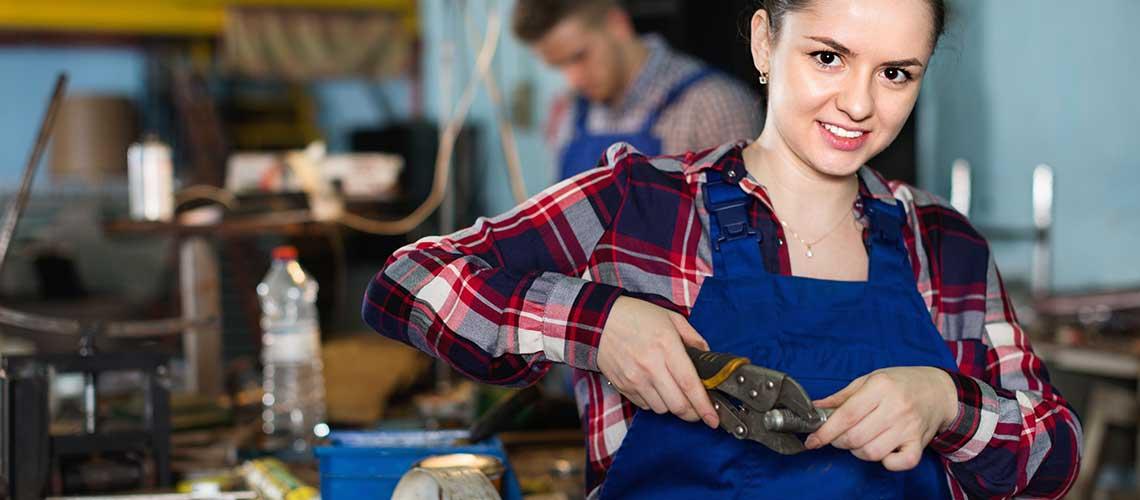 Jeune femme en bleu de travail manipulant un outil. Un jeune homme en arrière plan